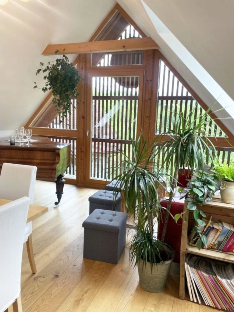Dachzimmer mit Giebelfenster, Pflanzen, Klavier und Sitzgelegenheiten.