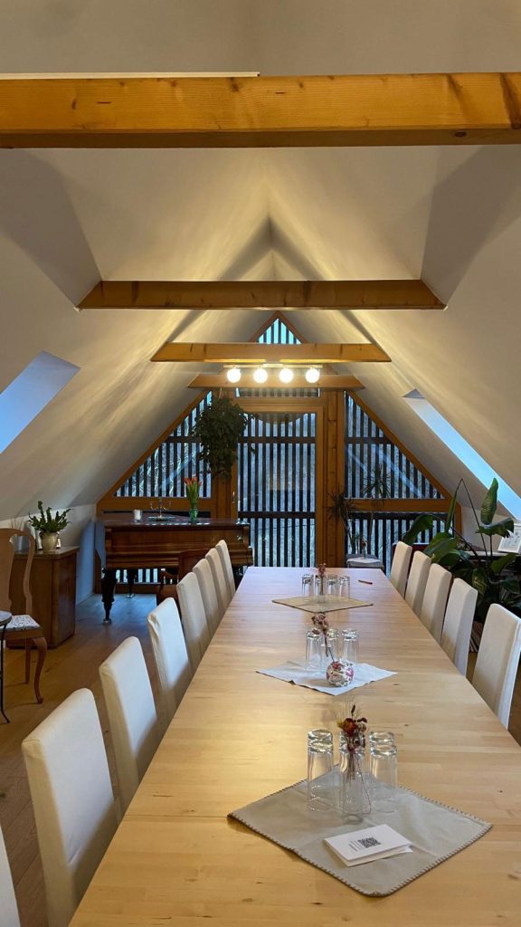 Ein langer Holztisch in einem hellen Raum im Dachgeschossstil mit schrägen Decken und Tageslicht.