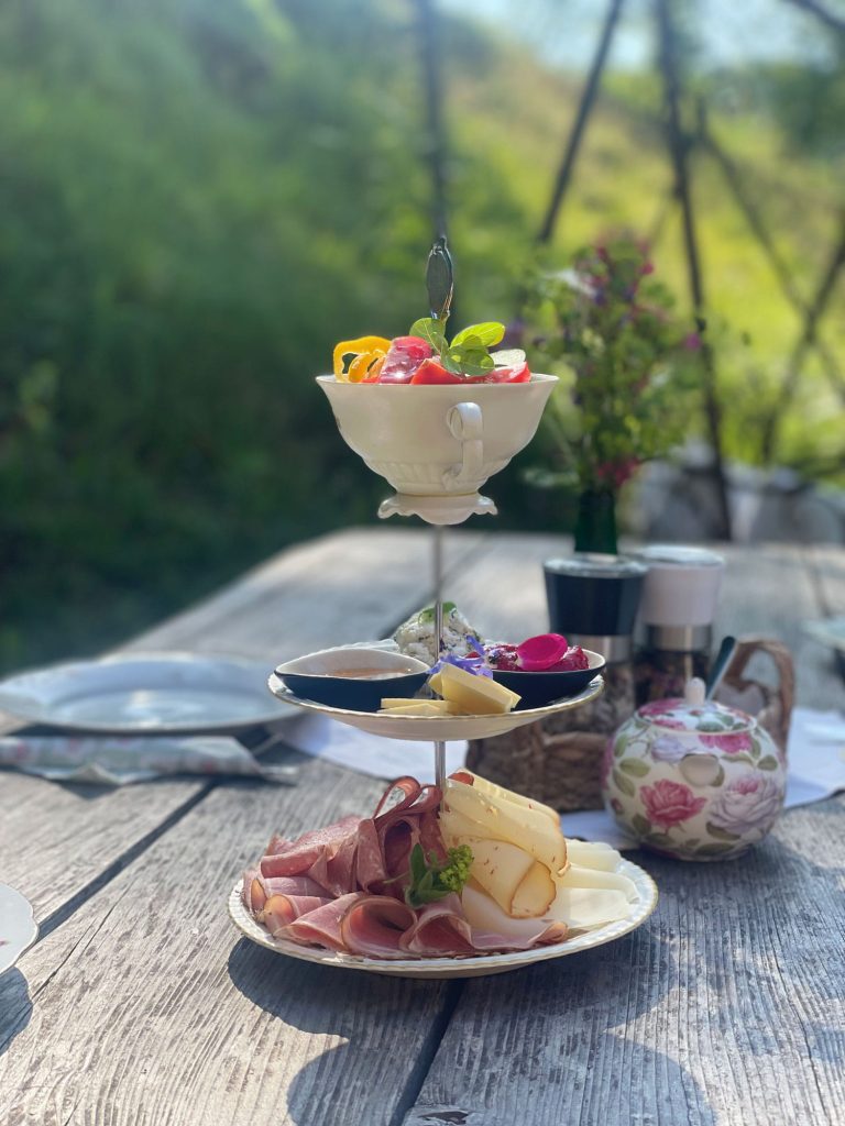Frühstücksetagere mit Wurstwaren, Käse und bunten Beilagen auf einem rustikalen Holztisch.
