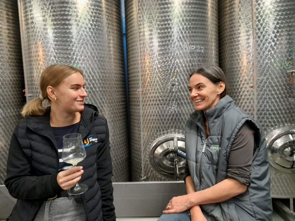 Zwei Frauen lächeln in einem Weingut und stehen vor großen Edelstahltanks.