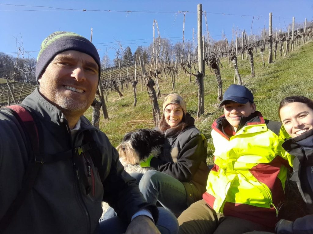 Eine Gruppe mit Hund genießt den Blick auf die Weinberge beim Weingut. Winterliche Weinberglandschaft, sonniger Tag.
